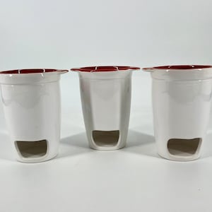 May include: Three white ceramic vases with a red rim and a rectangular opening near the base. The vases are cylindrical and have a glossy finish. The background is plain white.