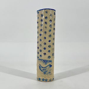 May include: A cylindrical ceramic vase with a cream-colored base and blue accents. The vase features a pattern of blue dots and two blue bird illustrations. The rim and base are also trimmed in blue.