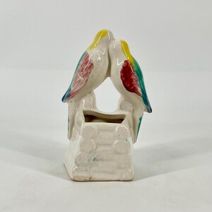 May include: A ceramic planter featuring two birds perched atop a structure resembling a log cabin. The birds are painted with yellow heads, red wings, and green accents, while the base is a glossy off-white with a textured, stacked log design.