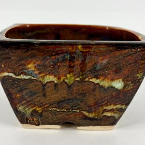 May include: A square ceramic bonsai pot with a tapered base and a glossy, multi-toned glaze featuring streaks of dark brown, amber, and pale green. The exterior surface has a textured, wood-like appearance with a small stamped mark near the bottom.