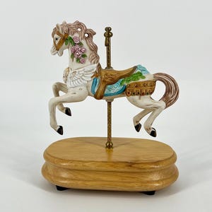 May include: A ceramic carousel horse figurine in white with a brown mane and tail, adorned with pink roses on its neck and white wings. It features a blue and gold saddle, mounted on a thin brass pole attached to an oval wooden base.