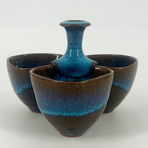 May include: A ceramic condiment server featuring three connected bowls arranged in a triangular formation. The exterior and interior surfaces display a deep brown base color with a vibrant, glossy turquoise blue glaze dripping from the center handle.