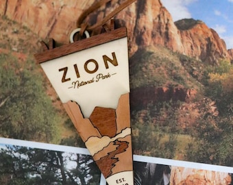 Peut inclure: Un ornement triangulaire en bois en forme de fanion représentant le parc national de Zion. La pièce présente le texte ZION National Park et EST. 1919, avec des tons de bois superposés illustrant des paysages de canyon et un cordon en cuir.