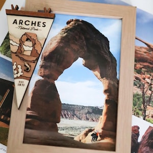May include: A light wood frame containing a photograph of the Arches National Park rock formation, accompanied by a triangular wooden pennant featuring the text ARCHES National Park and EST 1971 with a layered cutout of the rock arch.
