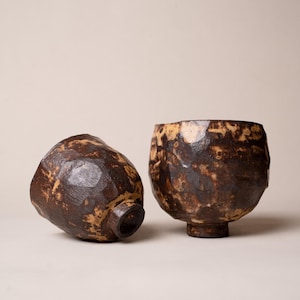 May include: Two handmade ceramic bowls with a rustic, textured appearance. The bowls are primarily dark brown with lighter tan accents, showcasing a unique, organic design. One bowl is upright, the other is on its side.