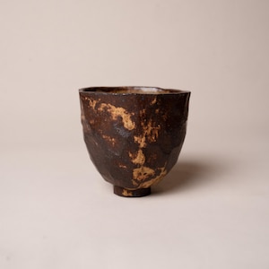 May include: A handcrafted, textured ceramic bowl with a dark brown exterior and lighter brown accents. The bowl has a unique, irregular shape and sits on a small, circular base. This rustic piece is ideal for serving or display.