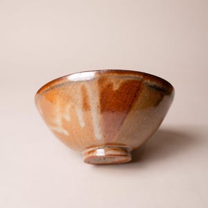May include: A ceramic bowl featuring a glossy glaze with blended tones of warm amber, deep brown, and cream. The bowl has a wide, circular opening, a tapered body, and a small, raised circular foot at the base, showcasing a smooth, reflective surface.