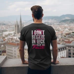 Peut inclure: T-shirt noir avec l'inscription "I DON'T BELIEVE IN LUCK SO I KEEP THE FAITH" en blanc et rose. La personne porte le t-shirt et regarde un paysage urbain.