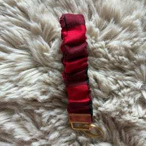 May include: A fabric wristlet keychain featuring a red and black buffalo plaid pattern. The fabric is gathered and ruched along a strap, attached to a gold-toned metal hardware clamp with an attached circular gold-toned metal key ring.