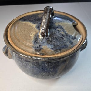 May include: A round ceramic lidded bowl featuring a mottled glaze in shades of deep blue, slate gray, and earthy tan. The bowl has two small side handles and a matching lid with a central arched handle and a small notch for a serving utensil.
