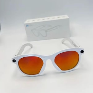 Puede incluir: Gafas de sol con montura blanca y lentes de color naranja vibrante. Las gafas de sol tienen una forma redondeada y un diseño elegante y moderno. Una caja blanca con un contorno de gafas de sol está en el fondo.