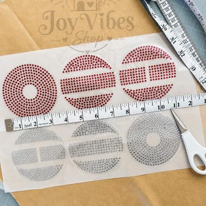 May include: A sheet of six circular iron-on rhinestone transfers featuring red and clear crystals in various geometric patterns. A white measuring tape shows the width of the transfer sheet is approximately 30.5 cm. A small pair of white scissors is included.
