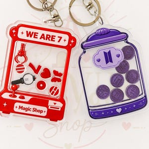 Pode incluir: Dois porta-chaves de acrílico com argolas prateadas. Um tem a forma de uma máquina de garra vermelha com o texto WE ARE 7 e Magic Shop, com berloques vermelhos. O outro é um frasco roxo com o logótipo BTS, contendo fichas circulares roxas.