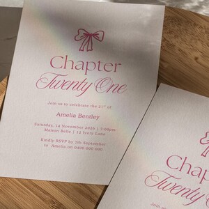 May include: Two off-white invitation cards with pink text and a bow illustration. The text reads "Chapter Twenty One" and includes details for a celebration, including a name, date, time, and RSVP information. The cards are on a wooden tray.