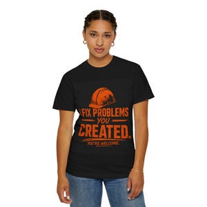 Puede incluir: Camiseta negra con un gráfico naranja de un casco de construcción y el texto "I FIX PROBLEMS YOU CREATED. YOU'RE WELCOME." La camiseta está hecha de un material suave.