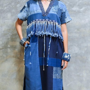 Può includere: Un abito blu patchwork al ginocchio con scollo a V rifinito da un bordo a motivi blu scuro, maniche corte e uno strato di frange decorative sul petto. Il capo è realizzato con vari pannelli di tessuto in diverse tonalità di blu.