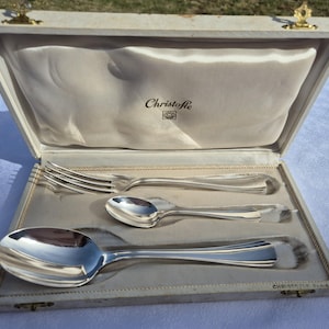 Peut inclure: Un ensemble de couverts argentés Christofle comprenant une fourchette, une petite cuillère et une grande cuillère, présenté dans un coffret de rangement crème à charnières avec un intérieur en satin portant le nom et le logo Christofle.