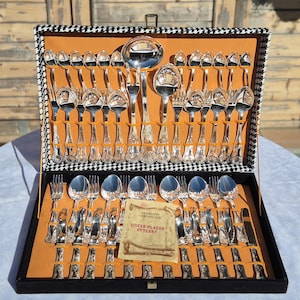 May include: A silver-plated cutlery set housed in a black and white houndstooth patterned case with an orange interior. The set includes various spoons, forks, and a ladle, all featuring ornate handles. A certificate of authenticity is included.