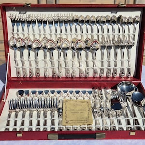 May include: A large silver-plated flatware set housed in a red carrying case. The set includes numerous forks, spoons, and knives arranged in rows, along with a serving ladle and a small certificate card with the text Silver plated cutlery set.