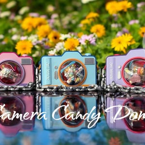 May include: Three colorful camera-shaped candy domes in pink, blue, and purple, each with a clear lens revealing colorful candies. The cameras are linked by a silver chain and sit on a reflective surface, with a floral background.