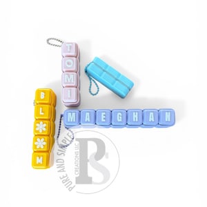May include: Four modular keychains made of square plastic blocks in yellow, light purple, and blue. One yellow keychain spells BL with two flower icons and M. One purple keychain spells TOMI. One blue keychain spells MAEGHAN. One blue keychain is blank.