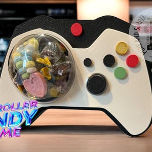 May include: A white and black video game controller shaped candy dish with a clear dome filled with colorful candies. The text "CONTROLLER CANDY DOME" is printed on the front of the dish.