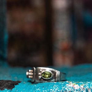 May include: A silver ring with a green oval gemstone. The ring has a rectangular design with decorative elements on the sides. The ring is set against a blue and black textured background.