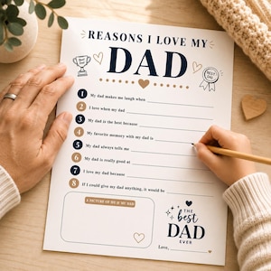 May include: A white paper with the text "REASONS I LOVE MY DAD" and fill-in-the-blank prompts. The paper includes decorative hearts, a trophy, and a ribbon. A hand is writing on the paper with a pencil.
