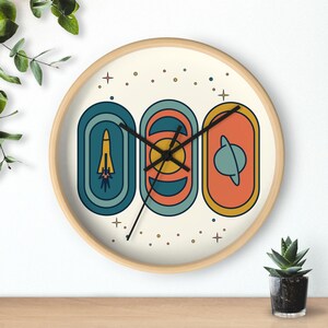 May include: A round wall clock with a light wood frame. The clock face features three retro-style, vertical oval designs in teal, orange, and yellow, depicting a rocket, sun, and planet. The clock hands are black.