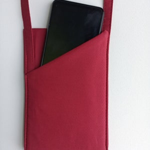 May include: A deep red fabric phone pouch with a diagonal pocket opening. The pouch features a matching red adjustable strap with a black plastic buckle at the top. A black smartphone is partially inserted into the pouch, showing its dark screen.