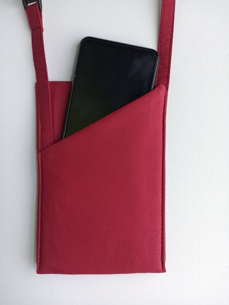 May include: A deep red fabric phone pouch with a diagonal pocket opening. The pouch features a matching red adjustable strap with a black plastic buckle at the top. A black smartphone is partially inserted into the pouch, showing its dark screen.