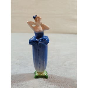 Può includere: Una statuetta in ceramica di una persona con busto e testa color pesca, che indossa un corpetto blu scuro e una gonna a forma di fiore blu. La base è verde e a forma di foglie, con un piccolo dettaglio a fiocco blu tra i capelli.