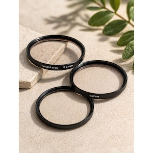 May include: Three black Quantaray 52mm camera lens filters. One filter is labeled "Quantaray 52mm". The other filter is labeled "JAPAN". The filters are arranged on a light beige surface with a small stone and green leaves.