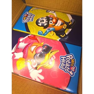 Könnte beinhalten: Zwei Mr. und Mrs. Potato Head Spielzeugboxen in einem Karton. Die Mr. Potato Head Box hat ein Piraten-Design mit schwarzem Hut und orangefarbener Jacke. Die Mrs. Potato Head Box hat ein rosa und gelbes Farbschema.