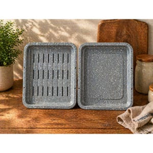 May include: Two gray speckled rectangular baking pans. One pan has vertical slots, the other is solid. The pans are on a wooden surface with a cutting board, jars, and a plant in the background. The pans are likely for baking or roasting.