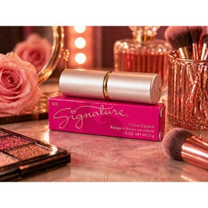 May include: A silver lipstick tube with gold accents rests on a pink box. The box reads "Signature" in cursive and "Creme Lipstick" with the weight of .13 oz. The background includes makeup brushes and a rose.