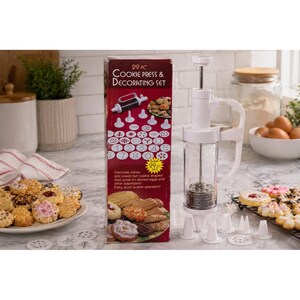 Könnte beinhalten: Ein weißes Keks- und Dekorationsset mit einem transparenten Zylinder und verschiedenen Spitzformen. Auf der Box steht "29 PC Cookie Press & Decorating Set" und zeigt Beispiele für Kekse. Ein Teller mit dekorierten Keksen befindet sich im Vordergrund.