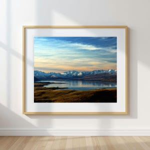 May include: A framed landscape print featuring a serene mountain lake scene. The artwork showcases snow-capped mountains under a blue and orange sky, with the lake reflecting the colors. The foreground displays golden-brown grassy fields.