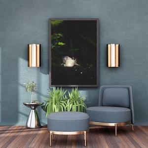 May include: A modern living room scene with a framed artwork, a blue armchair, and a matching ottoman. The room features a silver side table with a plant, two wall sconces, and a wooden floor.