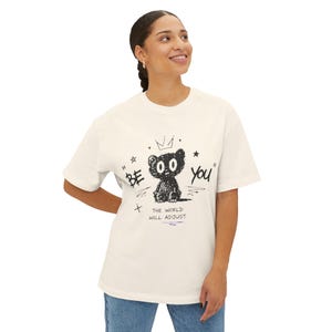 以下が含まれることがあります： オフホワイトの半袖Tシャツ。前面には、王冠をかぶった小さなクマ、星、そして「BE YOU」「THE WORLD WILL ADJUST」という文字が黒いグラフィックでプリントされています。デザインは手描き風のスケッチのような仕上がりです。