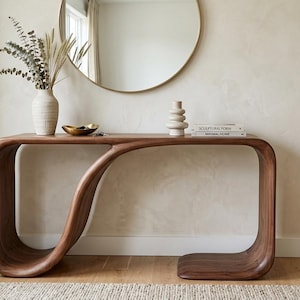 Op de afbeelding: Een moderne console met een uniek, vloeiend ontwerp van donker hout. De tafel heeft een gebogen basis en bovenkant, met een centrale golfachtige steun. Boven de tafel hangt een ronde gouden spiegel, die is versierd met een vaas, een kom en boeken.