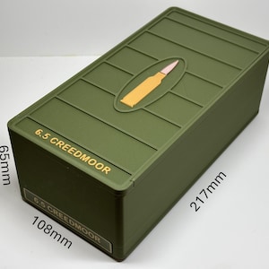 May include: An olive green rectangular storage box with a raised oval design featuring a yellow bullet. The box is labelled "6.5 CREEDMOOR" and measures 21.7cm long, 10.8cm wide, and 6.5cm high.