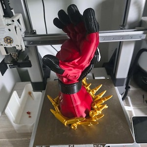 May include: A 3D printed sculpture of a red and black glove with a black wrist cuff, surrounded at the base by a ring of gold-colored bullet-shaped objects. The sculpture is positioned on the metal print bed of a 3D printer with visible components.