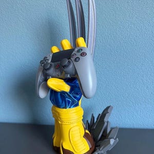 May include: A yellow and blue glove-shaped controller stand featuring three silver-colored blades extending upward from the knuckles. The stand holds a grey PlayStation controller and includes a jagged grey base resembling broken rock fragments.