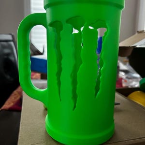 May include: A bright green mug with a large handle and a cylindrical body. The mug features a cut-out design of the Monster Energy logo. The mug is made of a solid material and is sitting on a cardboard surface.