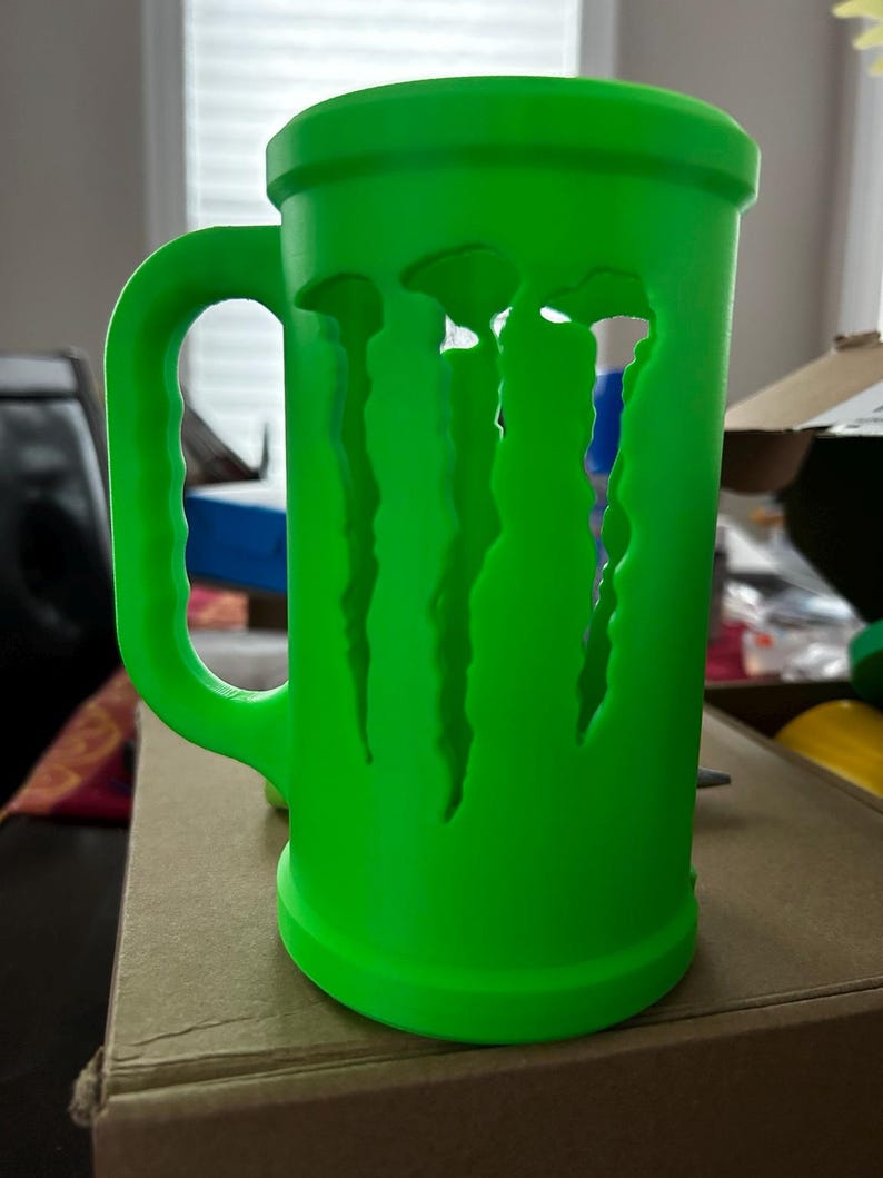 May include: A bright green mug with a large handle and a cylindrical body. The mug features a cut-out design of the Monster Energy logo. The mug is made of a solid material and is sitting on a cardboard surface.