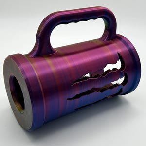 May include: A cylindrical object with a handle, featuring a color-shifting finish of purple, blue, and gold. The design includes claw-shaped cutouts along the side. The object has a circular opening on one end.