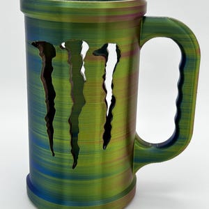 May include: A large, cylindrical mug with a handle, featuring a multi-colored, textured finish in shades of green, blue, and purple. The mug has a distinctive design with three claw-like cutouts on the front. The mug is designed to hold beverages.