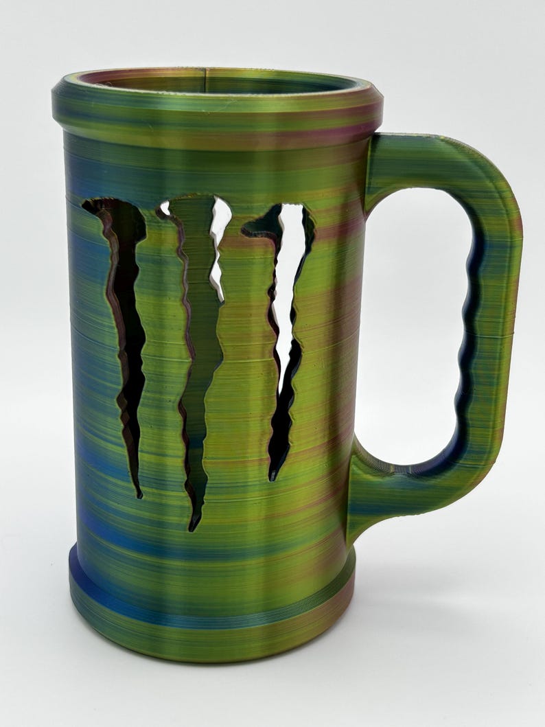May include: A large, cylindrical mug with a handle, featuring a multi-colored, textured finish in shades of green, blue, and purple. The mug has a distinctive design with three claw-like cutouts on the front. The mug is designed to hold beverages.