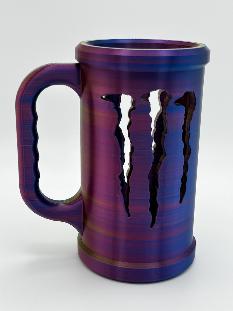 May include: A cylindrical mug with a large handle, featuring a color-shifting finish of purple, blue, and gold. The mug has three vertical, jagged cutouts on the side. The mug is designed to hold beverages.
